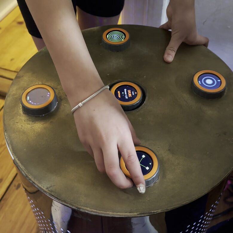 The built TAMBUM table seen from above: two hands playing on the round top, four discs in a circle and the master in the middle, all lit
