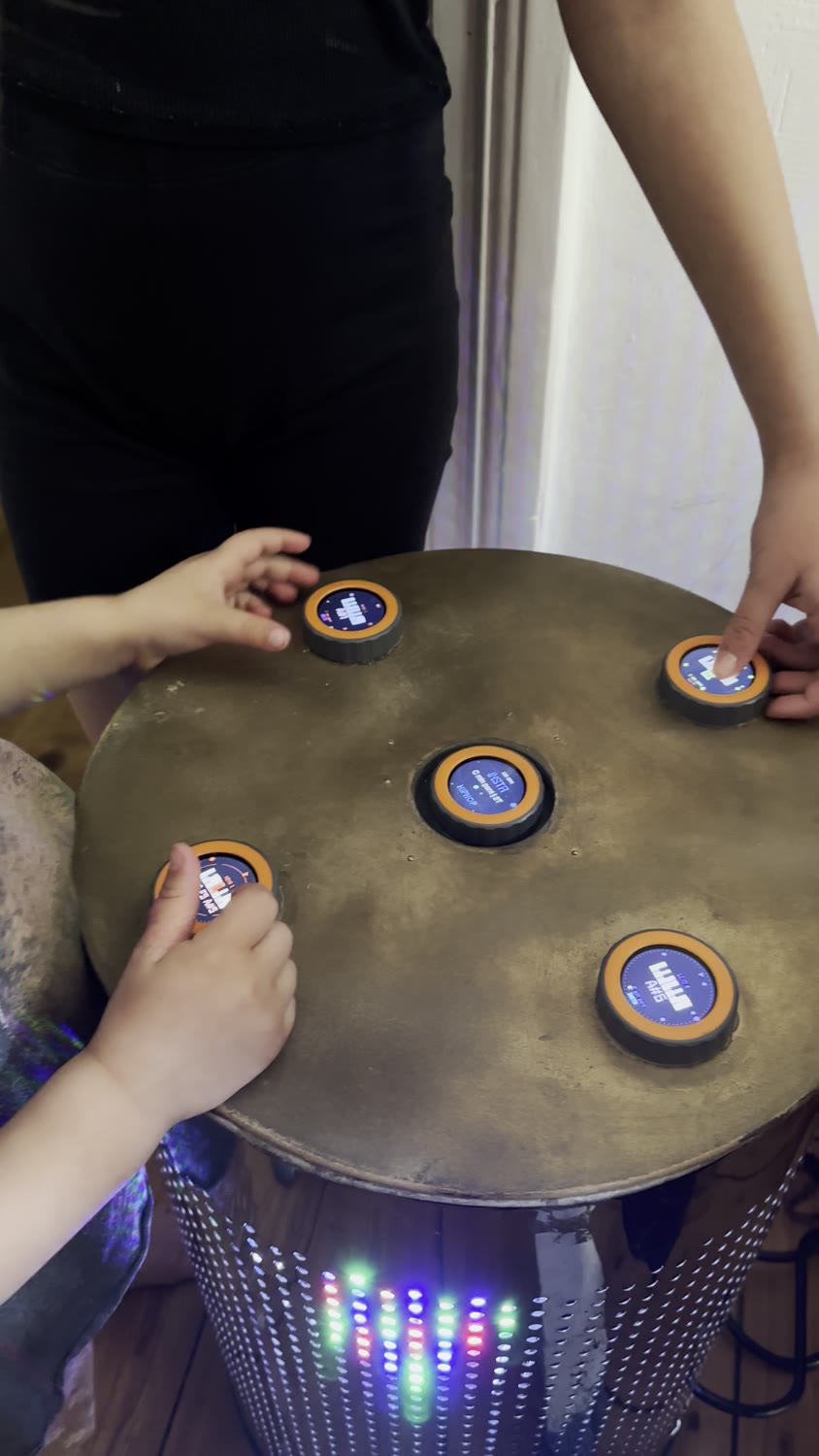 Two people playing together at the round TAMBUM table, coloured lights glowing in the base