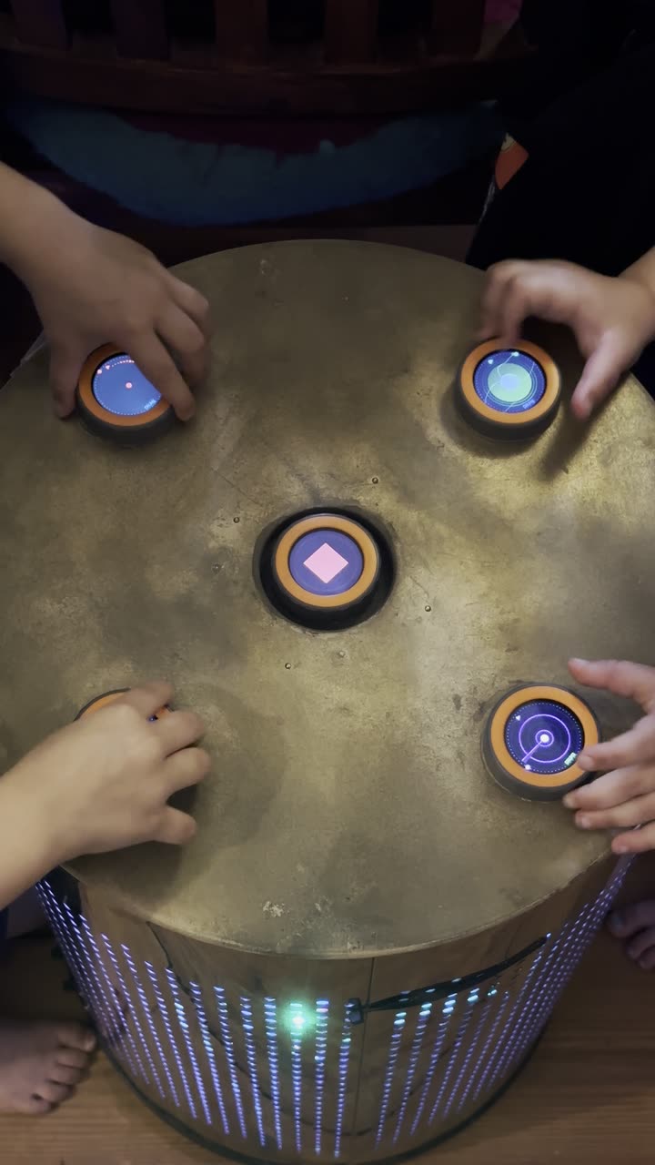 Seen from above: four children's hands reach in from every side for the discs of the TAMBUM table, the base glowing blue and green