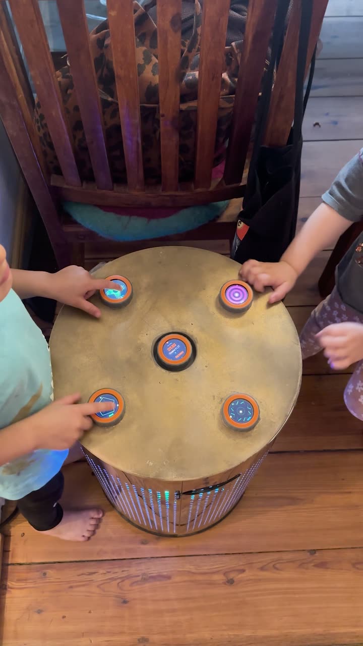 Seen from above: two children stand at the TAMBUM table reaching for different discs at the same time, all five lit, light running in the base