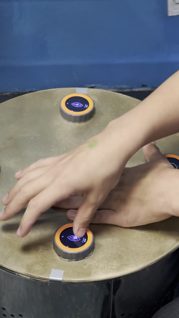 Two hands at the TAMBUM table: one finger presses a disc, a pattern showing on its display