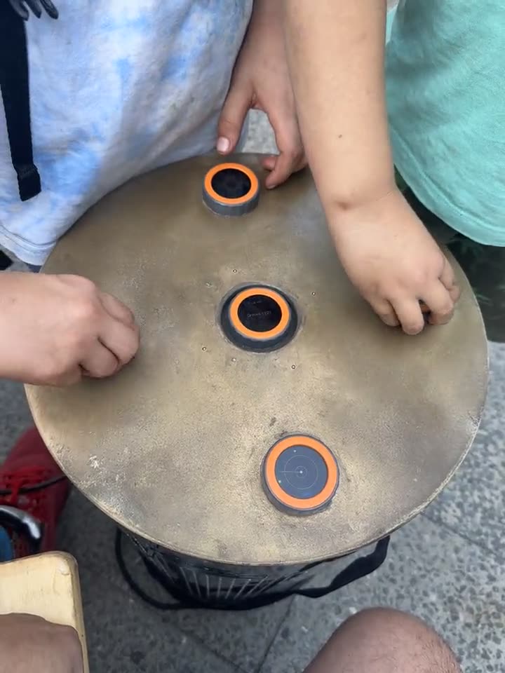 Top view of the round TAMBUM top outdoors: three discs with orange rings, several children’s hands reaching in at once from different sides